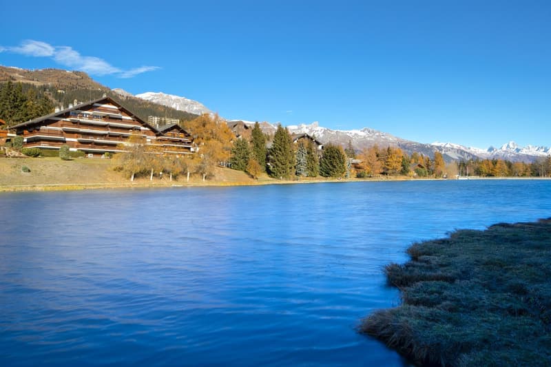 Large building with many windows, surrounded by trees, mountains, and a lake
