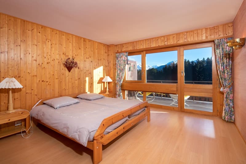 wooden bed, two pillows, wooden nightstand, lamp, wooden wall, large window, balcony, view of mountains