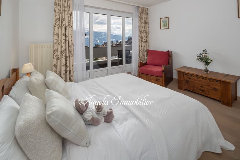 large bed, white bedding, pillows, wooden nightstand, lamp, radiator, curtains, picture frame, wooden chair, flower vase, wooden chest, balcony with chairs, scenic mountain view