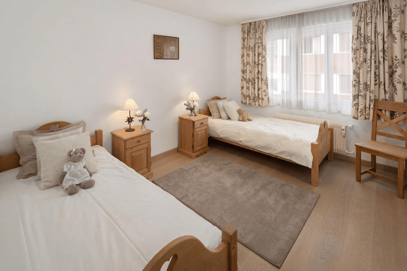 Two single beds, wooden nightstands, lamps, stuffed animals, rug, wooden chair, picture frame, white walls, white curtains