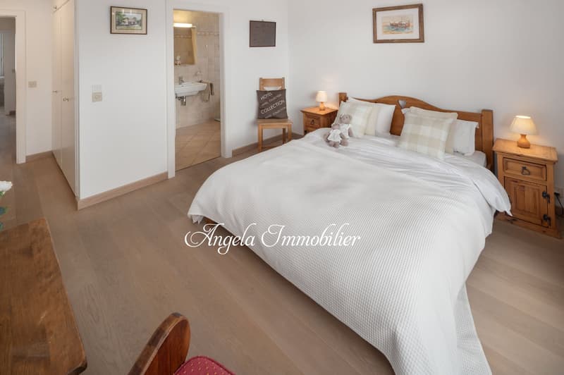 Bedroom with wooden floor, white walls, large bed, wooden nightstands, lamps, chair, and decorative items. En-suite bathroom with sink and mirror.