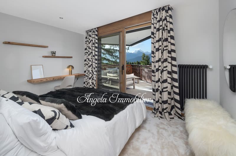 large bed, white bedding, black and white pillows, wooden shelves, desk, lamp, sliding glass door, balcony, mountain view
