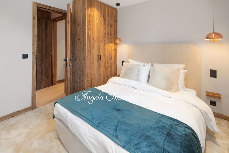 Bedroom with wooden door, wooden wardrobe, bed with white and blue bedding, beige headboard, wooden shelves, white walls, wooden floor, lamps