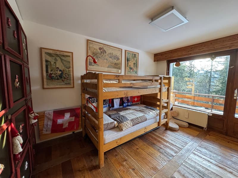A bedroom with wooden bunk beds, wooden floor, white walls, a radiator, a lamp, and a window with a view of the mountains.