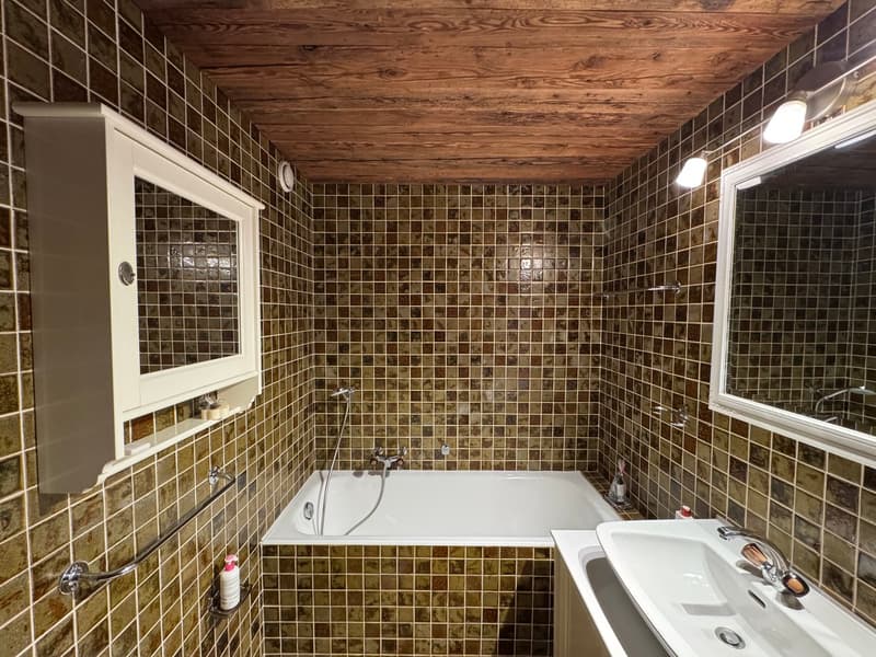 Bathroom with wooden ceiling, tiled walls, bathtub, sink, mirror, and a cabinet with a glass door.