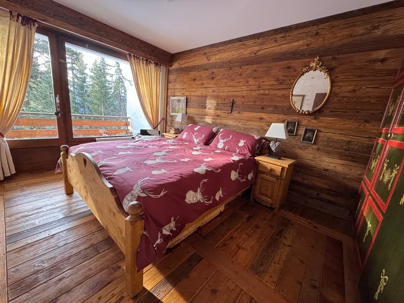 wooden bed, red bedspread, wooden nightstands, lamps, mirror, wooden walls, large window, curtains, picture frames