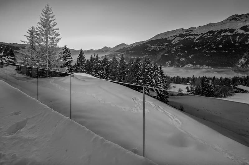 Snowy mountain landscape with pine trees, glass railing, and a distant view of mountains and a lake.