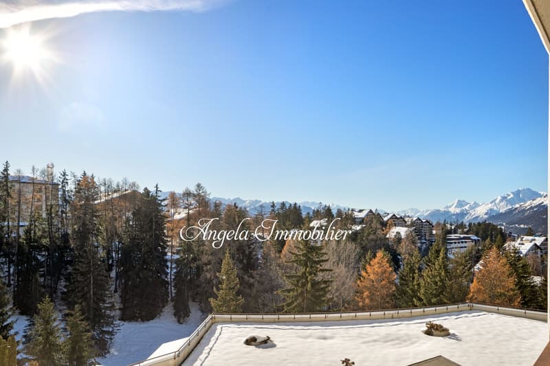 Wide view of snowy mountains, pine trees, houses, clear sky, and some footprints on the snow