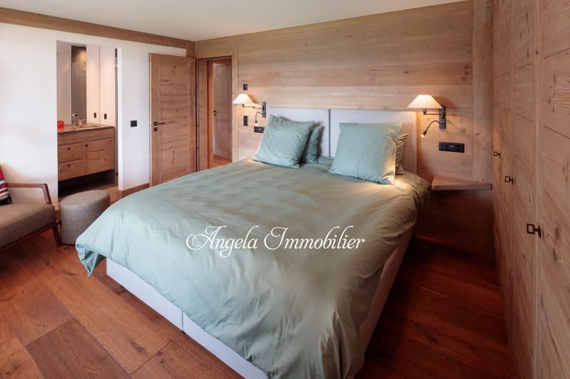 Spacious bedroom with wooden walls and floors, a large bed with light blue bedding, two bedside lamps, and a dresser in the adjacent room.