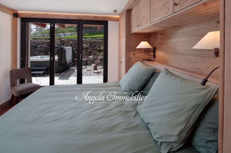 Spacious bedroom with wooden walls and floors, large windows overlooking a garden, a bed with light blue bedding, and a chair in the corner.