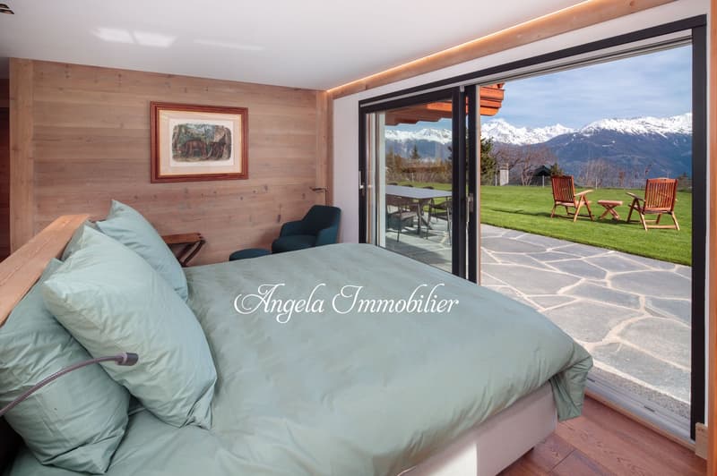 Spacious bedroom with wooden walls, large windows overlooking a scenic mountain landscape, a comfortable bed with green bedding, and a seating area with chairs facing the outdoor patio and view.