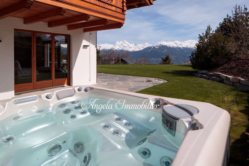 Wooden chalet-style house with large windows, surrounded by a grassy lawn and with a view of snow-capped mountains in the background. The house has a hot tub or jacuzzi on the patio overlooking the scenic landscape.
