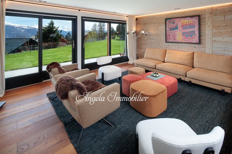 Spacious living room with large windows overlooking a grassy outdoor area and mountains in the background. The room features a comfortable sofa, armchairs, and ottomans in neutral tones, as well as a colorful abstract artwork on the wall. The room has a c