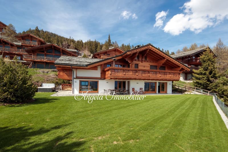 A group of traditional Swiss-style wooden chalets with sloped roofs, balconies, and large windows overlooking a well-manicured lawn. The chalets appear to be part of a resort or vacation community set in a forested mountain landscape.