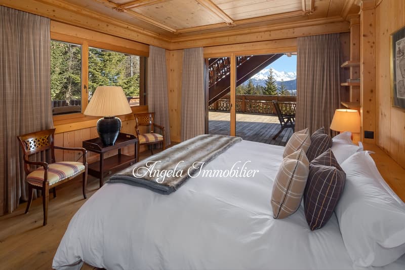 Spacious bedroom with wooden walls and ceiling, large windows overlooking a mountain landscape, a king-size bed with plush bedding, armchairs, and a table lamp