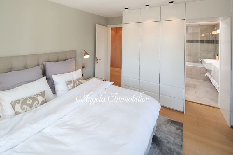 Bedroom with white bed, gray rug, gray headboard, several pillows, lamps, large white closets, wooden door to bathroom with bathtub.