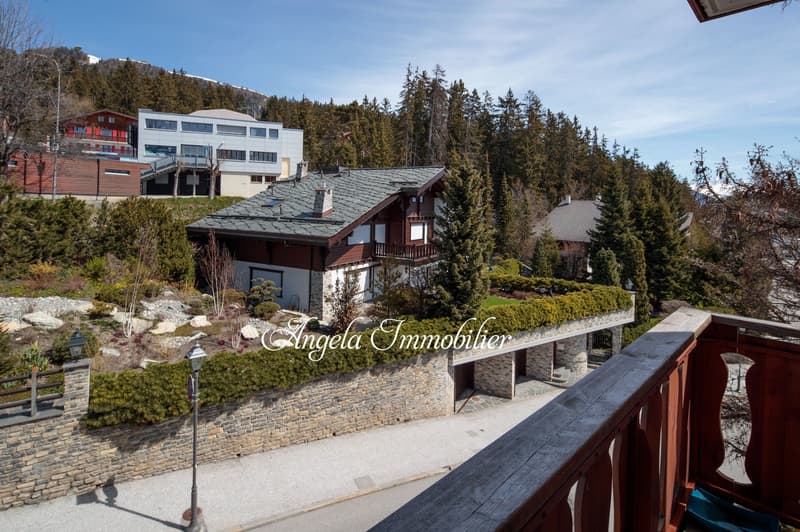 Wooden chalet style house, gray roof, balcony with wooden railing, stone wall, garden, trees, mountain view