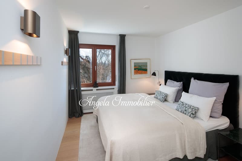 Contemporary style bedroom with white walls and wooden floor. A large double bed with a black headboard and multiple cushions. The window has wooden frame and gray curtains. Beside the bed is a side table with a lamp and a framed picture on the wall.