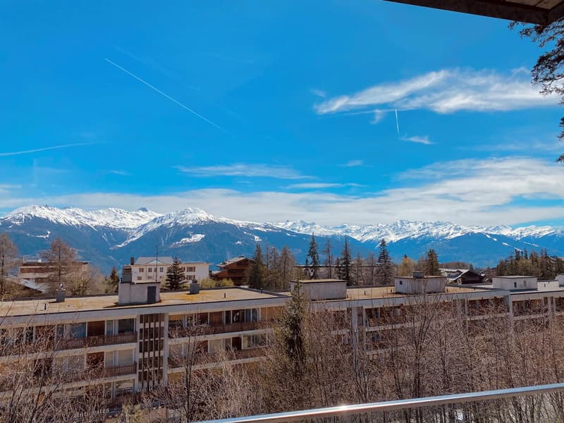 A multi-story building with balconies, surrounded by trees and mountains in the background