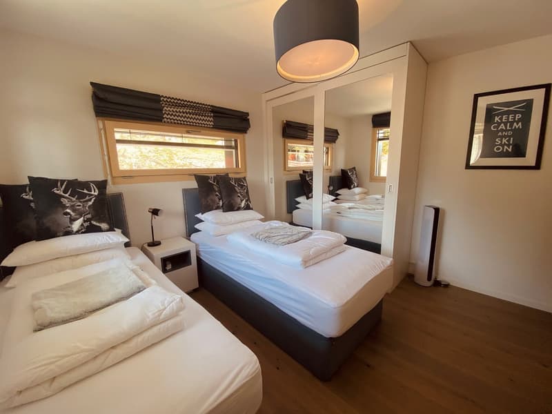 Bedroom with two single beds, white bedding, wooden floor, large windows, nightstands, lamps, mirror, picture frame, and a ceiling lamp.
