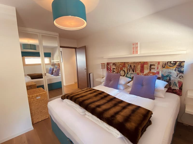 Bedroom with a bed, pillows, fur blanket, lamp, mirror, storage boxes, wooden floor
