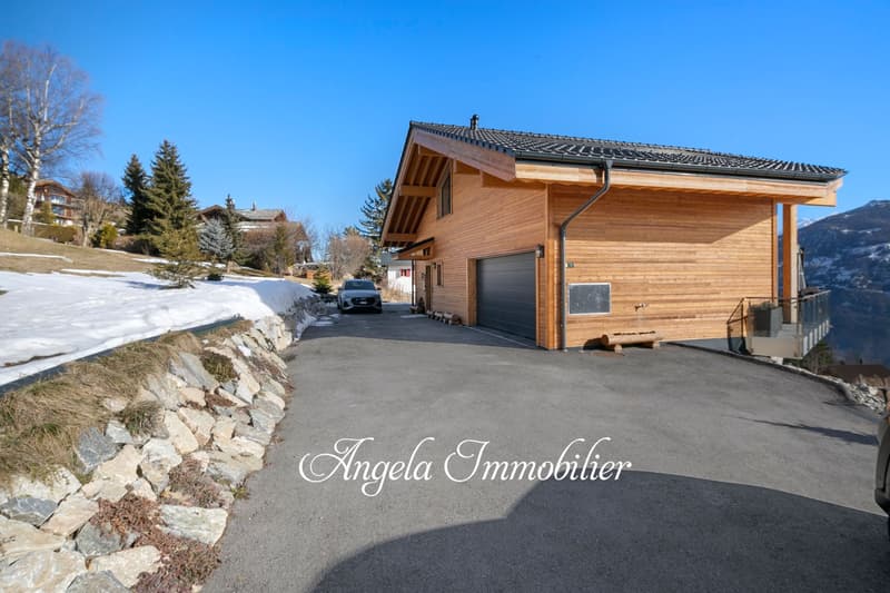 2-story wooden chalet-style house with a garage, surrounded by snow-covered trees and a paved driveway. The house has a tiled roof and a wooden exterior.