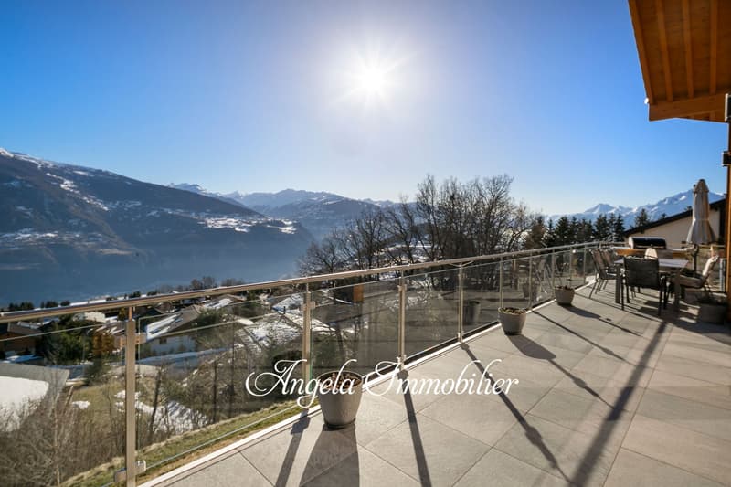 Large balcony with glass railing, overlooking a scenic mountain landscape with snow-capped peaks and a lake in the distance. The balcony has outdoor furniture and a potted plant, providing a comfortable space to enjoy the stunning views.