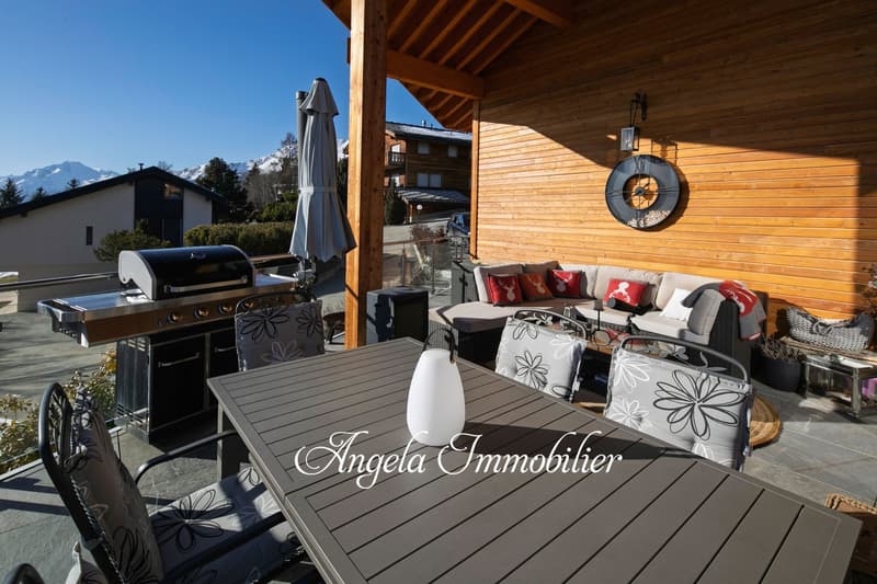 Wooden deck with outdoor furniture, including a sofa and chairs, a barbecue grill, and a table with the 'Angela Immobilier' logo. The deck is surrounded by a wooden structure with a slanted roof, and there are mountains visible in the background.