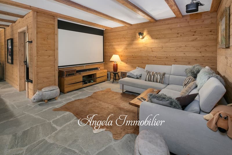 Cozy living room with wooden walls and ceiling, large gray sofa, coffee table, entertainment center with TV, and a fireplace. The room has a warm and rustic feel with the use of natural materials and neutral tones.