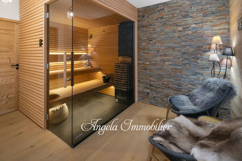 The image shows a cozy and luxurious spa-like room with a sauna, a stone accent wall, and a comfortable chair. The sauna has a glass door and wooden interior, and there is a fireplace visible in the room as well.