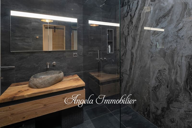 Luxurious bathroom with black marble tiles, a wooden vanity with a stone sink, and a sliding wooden door. The bathroom features modern lighting fixtures and a spacious shower area.