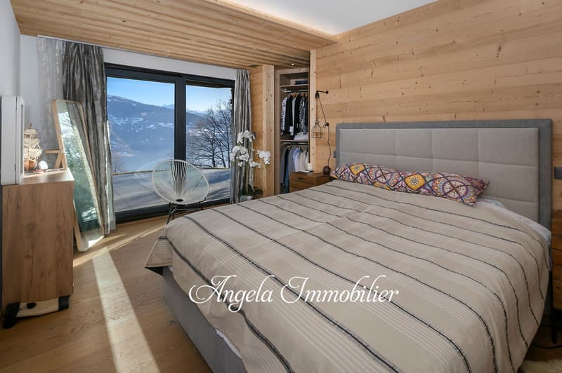 Spacious bedroom with wooden walls and ceiling, large windows offering a scenic mountain view, a comfortable bed with patterned bedding, a chair, and a closet for storage.