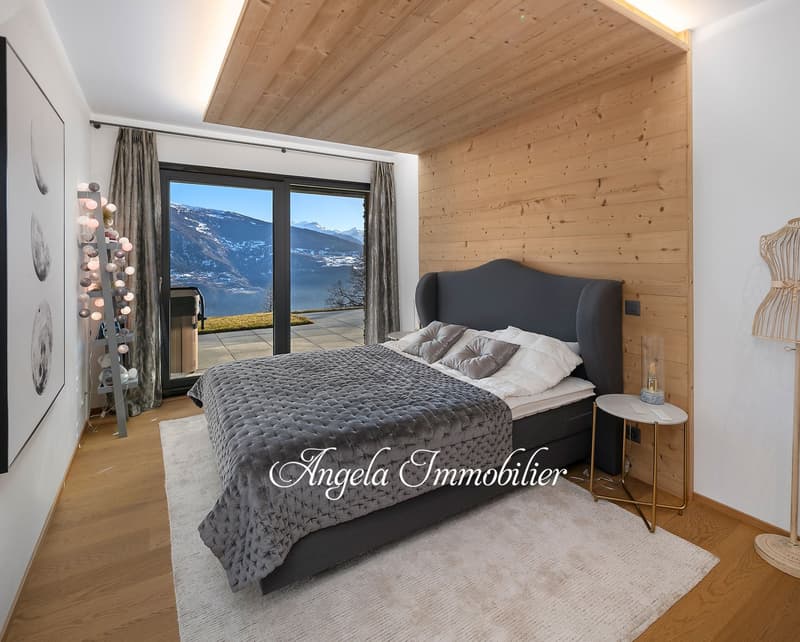 Spacious bedroom with wooden ceiling, large windows offering a scenic view, a plush gray bed with a quilted comforter, and side tables with lamps.