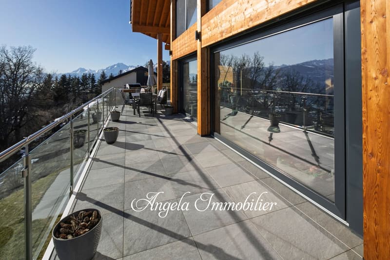 Large balcony or terrace with glass railing, wooden beams, and a view of mountains in the distance. The balcony has outdoor furniture and potted plants.