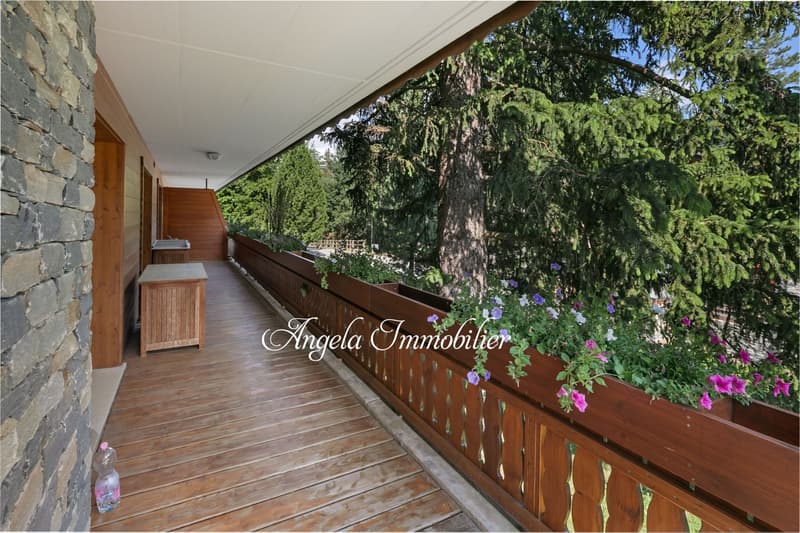 Covered wooden balcony or porch with stone walls, overlooking a lush, forested area with tall trees and flowering plants.