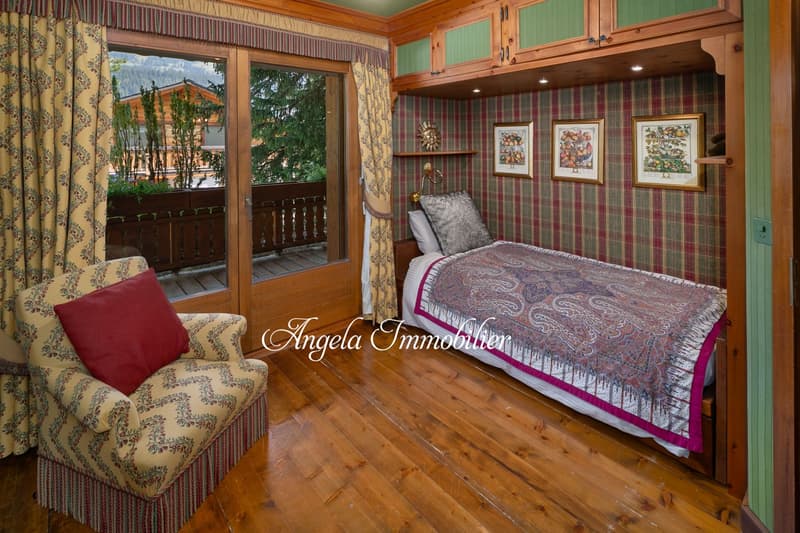 Cozy bedroom with wooden walls and floors, large windows overlooking a balcony, plaid wallpaper, floral curtains, armchair, and a patterned bedspread