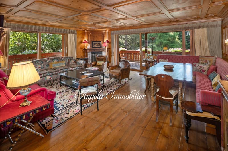 Spacious living room with wooden ceiling, large windows overlooking a balcony, ornate fireplace, floral patterned sofa, wooden dining table, and various antique furniture pieces