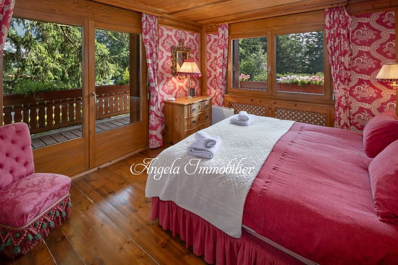 Spacious bedroom with wooden walls and floors, large windows overlooking a balcony and lush greenery outside, red and white patterned curtains, wooden dresser, and a cozy bed with a white comforter and red bedskirt