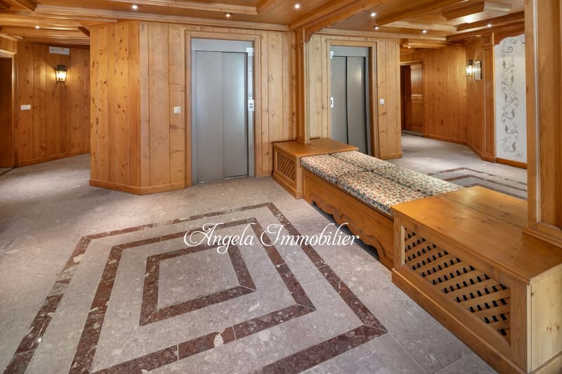 Spacious lobby area with wooden paneled walls, hardwood floors, decorative tile flooring, built-in bench seating, and an elevator. The space has a cozy and rustic feel with warm lighting and a fireplace visible.