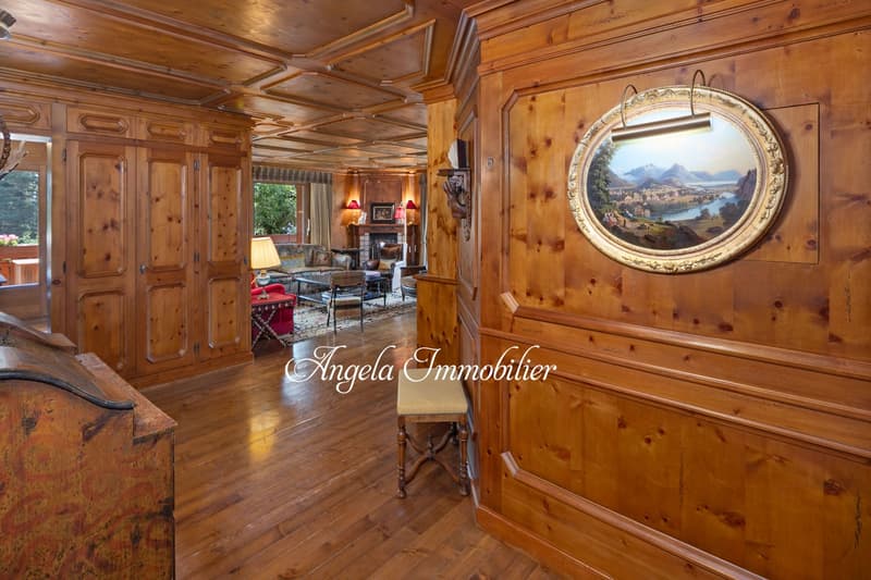 Spacious living room with wooden paneled walls and ceiling, hardwood floors, large ornate framed mirror, antique furniture, and a fireplace.