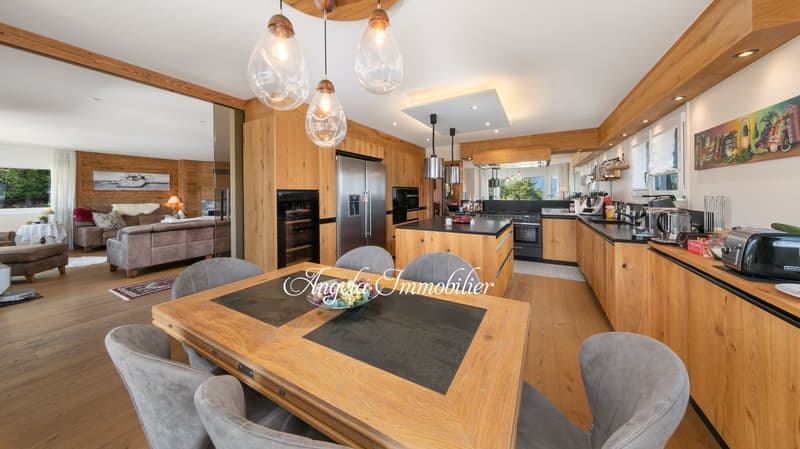 Spacious open-concept kitchen and living room with wooden walls and ceiling, large windows, modern appliances including a dishwasher, dining table with chairs, and a cozy living room area with a sofa and armchair.