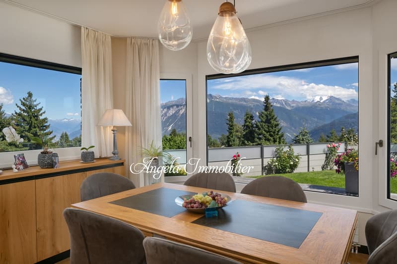 Spacious dining room with large windows offering a panoramic view of the mountains, wooden dining table with chairs, wooden sideboard, pendant lighting fixtures, and potted plants.