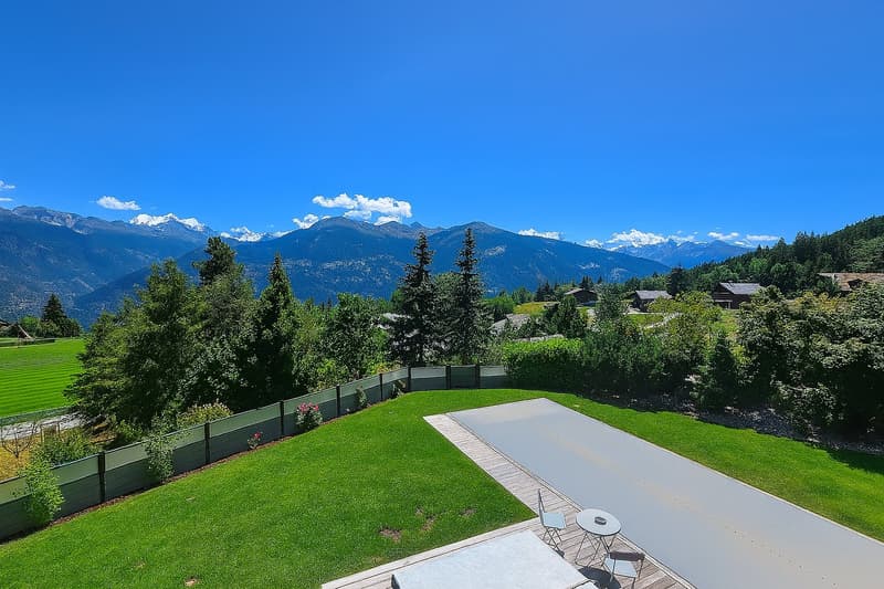 Large property with a swimming pool, surrounded by lush green trees and mountains in the background. The property has a well-manicured lawn and a wooden deck or patio area next to the pool.