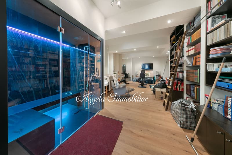 Large open-plan room with hardwood flooring, white walls, and built-in bookshelves. There is a blue-tinted glass wall or partition that appears to be a feature. The room contains various furniture and office equipment, suggesting it is used as a workspace