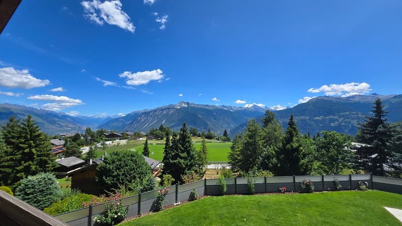 The image shows a scenic mountain landscape with a blue sky, fluffy white clouds, and lush green trees and vegetation. In the foreground, there is a well-manicured lawn and a wooden fence. In the background, there are snow-capped mountains and a picturesq