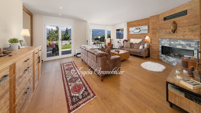Spacious living room with hardwood floors, wooden accent walls, large windows overlooking a garden, leather sofas, coffee table, and a stone fireplace