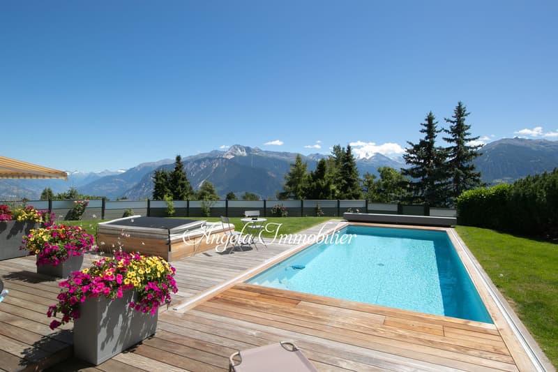 Outdoor swimming pool with wooden deck, surrounded by lush greenery and mountains in the background. The property also features a hot tub and beautiful flower planters.