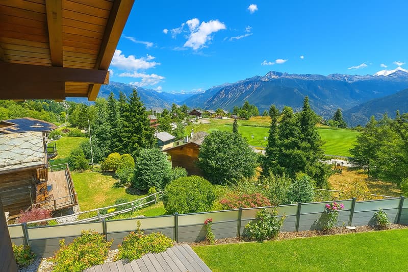 Wooden chalet-style house with a balcony overlooking a scenic mountain landscape. The house is surrounded by lush greenery, including trees, shrubs, and a well-manicured lawn. The view from the balcony showcases the majestic mountains in the distance unde