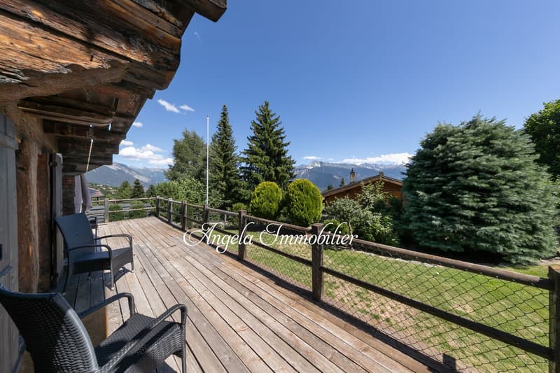 Wooden chalet-style house with a large wooden deck overlooking a scenic mountain landscape with pine trees and a clear blue sky. The deck has outdoor furniture and a railing with a wire mesh fence.