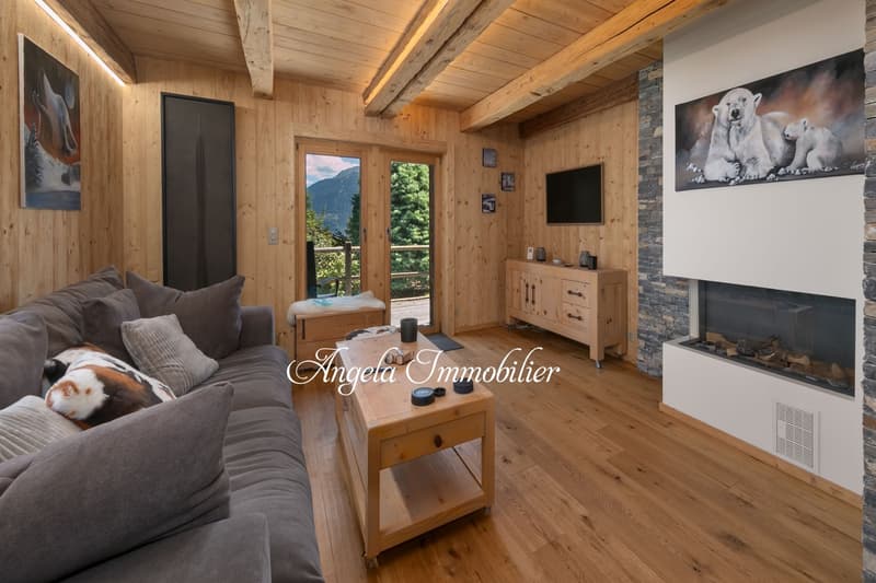Cozy living room with wooden ceiling and walls, large windows providing a scenic view, a fireplace, a gray sectional sofa, and a wooden coffee table and cabinet.